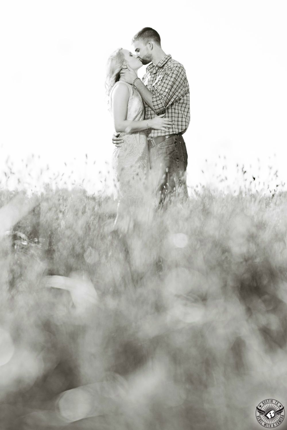 Flirty blond  girl wearing a light colored dress  is kissed and held tenderly by a dark haired guy wearing a black and white plaid long sleeve shirt and dark jeans in a field of golden wheat against a big Texas sky in this soft engagement photograph near Austin.